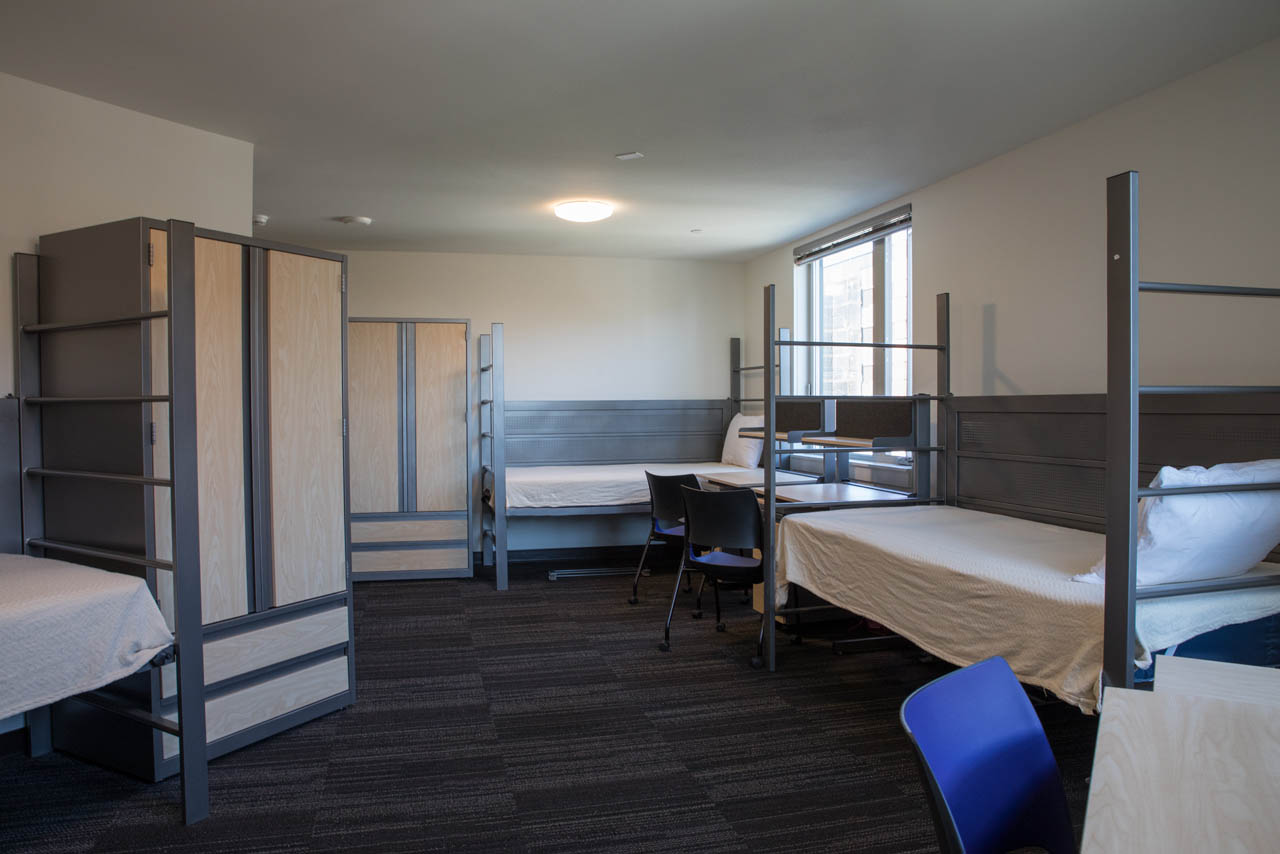 A dorm room with bunk beds, dressers, chairs, and desks.