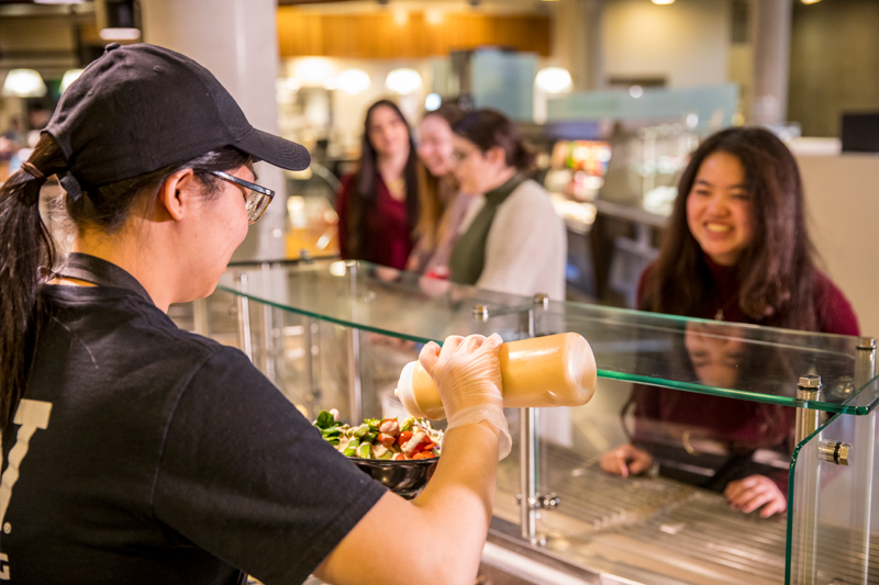 A Student working at Local Point dining hall pours on a bowl while customer smiles.
