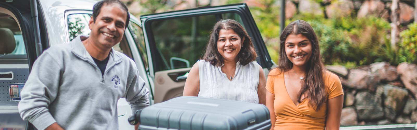 A student smiling with her parents in front of their car.