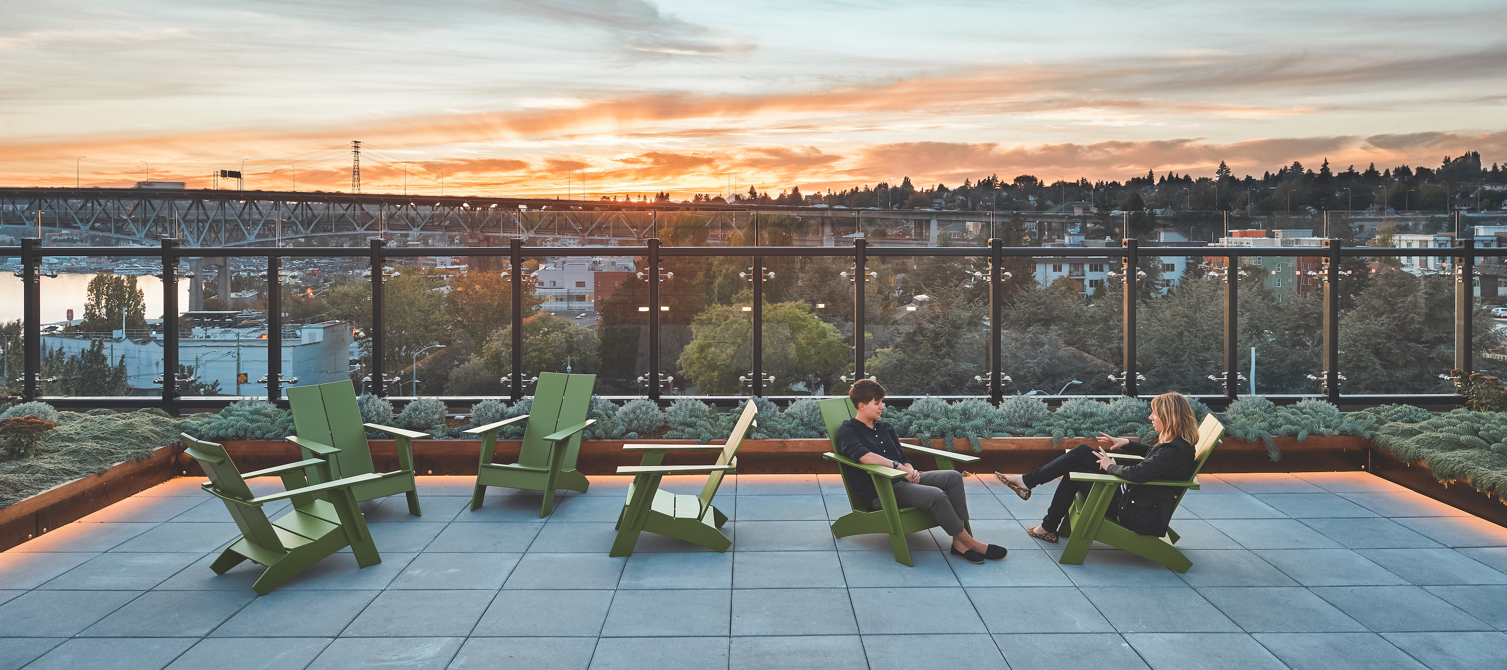 Two people sitting in green chairs on Terry Hall rooftop deck in front of sunset.