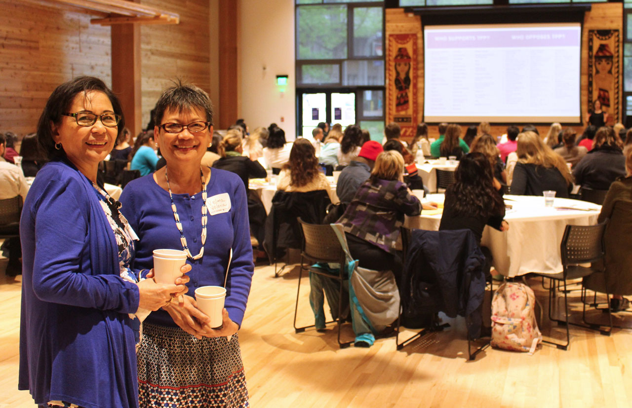 Two women smiling holding cups in a room of people sitting at tables and chairs.