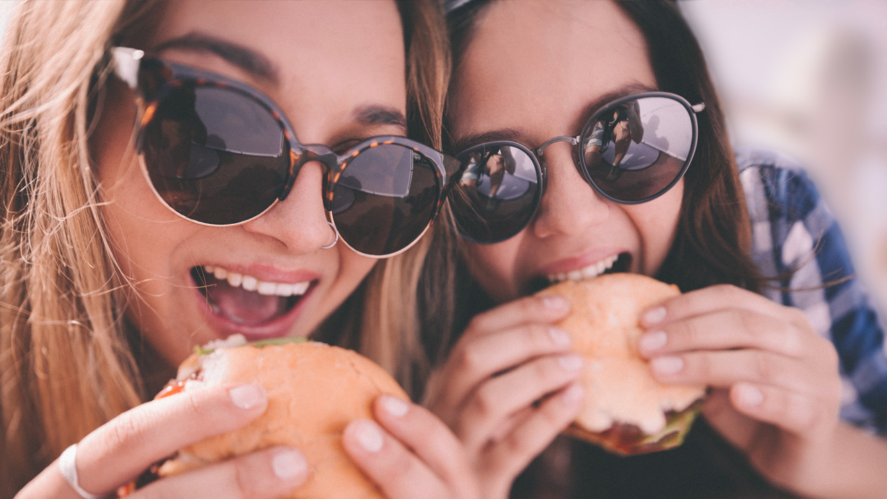 Two students wearing sunglasses take a bite of sandwiches.
