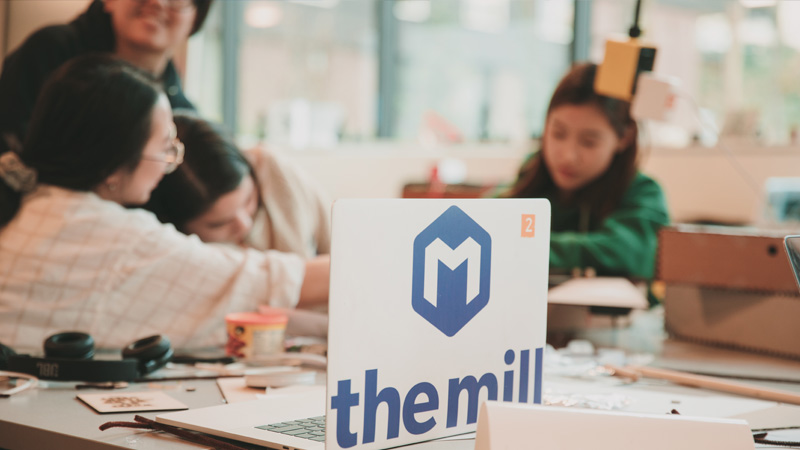 A laptop with a sticker reading "the mill" in front of students.
