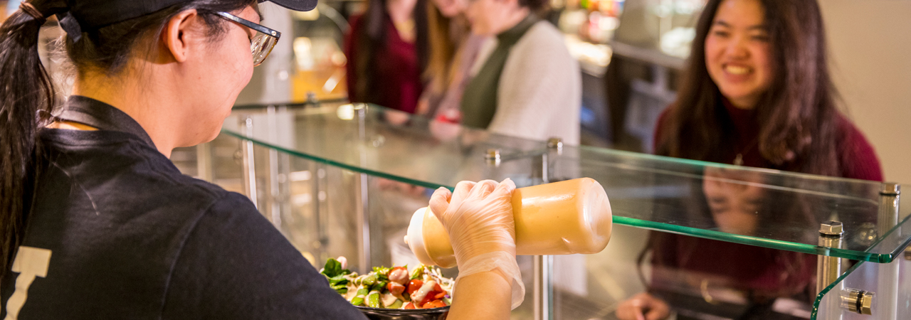 A student employee in Local Point pours sauce on bowl while student watches smiling.