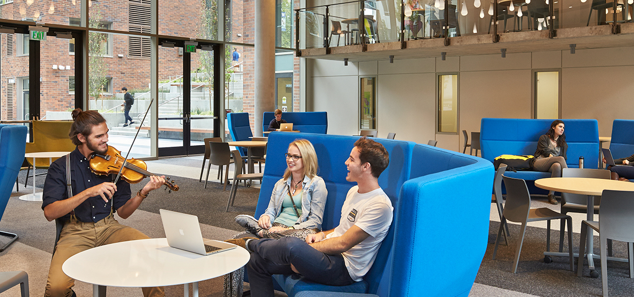 A student playing the violin to two people sitting on a blue couch in the Maple Great Room.