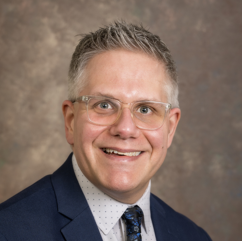 Phil Badaszewski smiking in front of gray background.