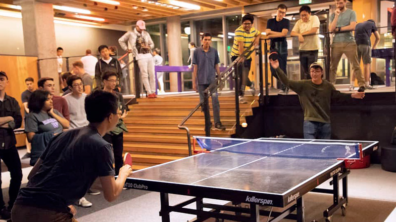 Students playing ping pong while a group of students watch in Area 01.