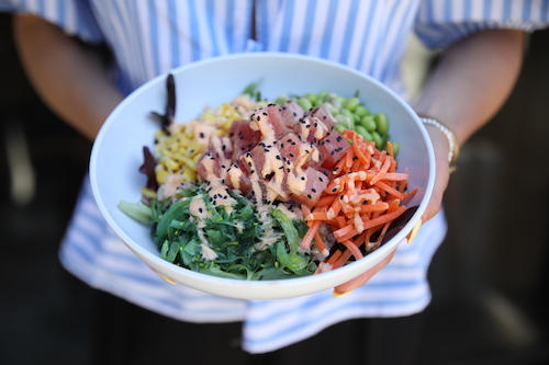A person in a blue striped shirt holding a bowl filled with poke, carrots, corn, edamame, and greens.
