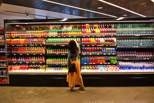 A woman picks a drink from refrigerated shelves in a grocery store. 